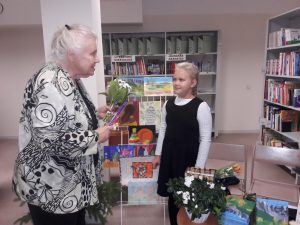 Krāsains foto. Bibliotēkas telpā vecā sieviete apsveic meiteni ar puķēm un vēl kaut ko