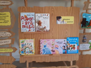 Krāsains foto. Uz koka statīvā saliktās dažādas bērnu grāmatas. Uzrakst uz lapas "Mazajiem pavāriem", blakus meitenes foto