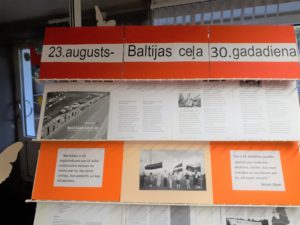 Krāsains foto. Uz dzelzs statīva vairākas rindās saliktās krāsainās lapas uz tiem izdrukātas lapas ar foto un tekstu. Augšā virsraksts - "23.augusts- Baltijas ceļa 30. gadadiena"