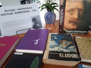 Krāsains foto. Tuvplāns, uz galda saliktās dažādās grāmatas, uz lapas uzraksts- "Norvēģu rakstniekam Knutam Hamsunam -160"