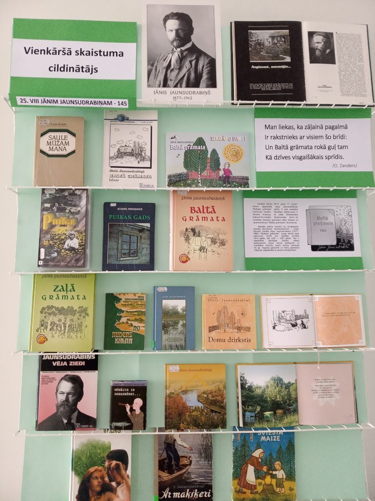 grāmatu kolekcija, kas izkārtota rindās uz zaļa fona. Kolekcijā ir dažādi nosaukumi un autori, un dažas grāmatas izskatās kā vecāki izdevumi. Ir arī izdrukātās lapas ar tekstu.