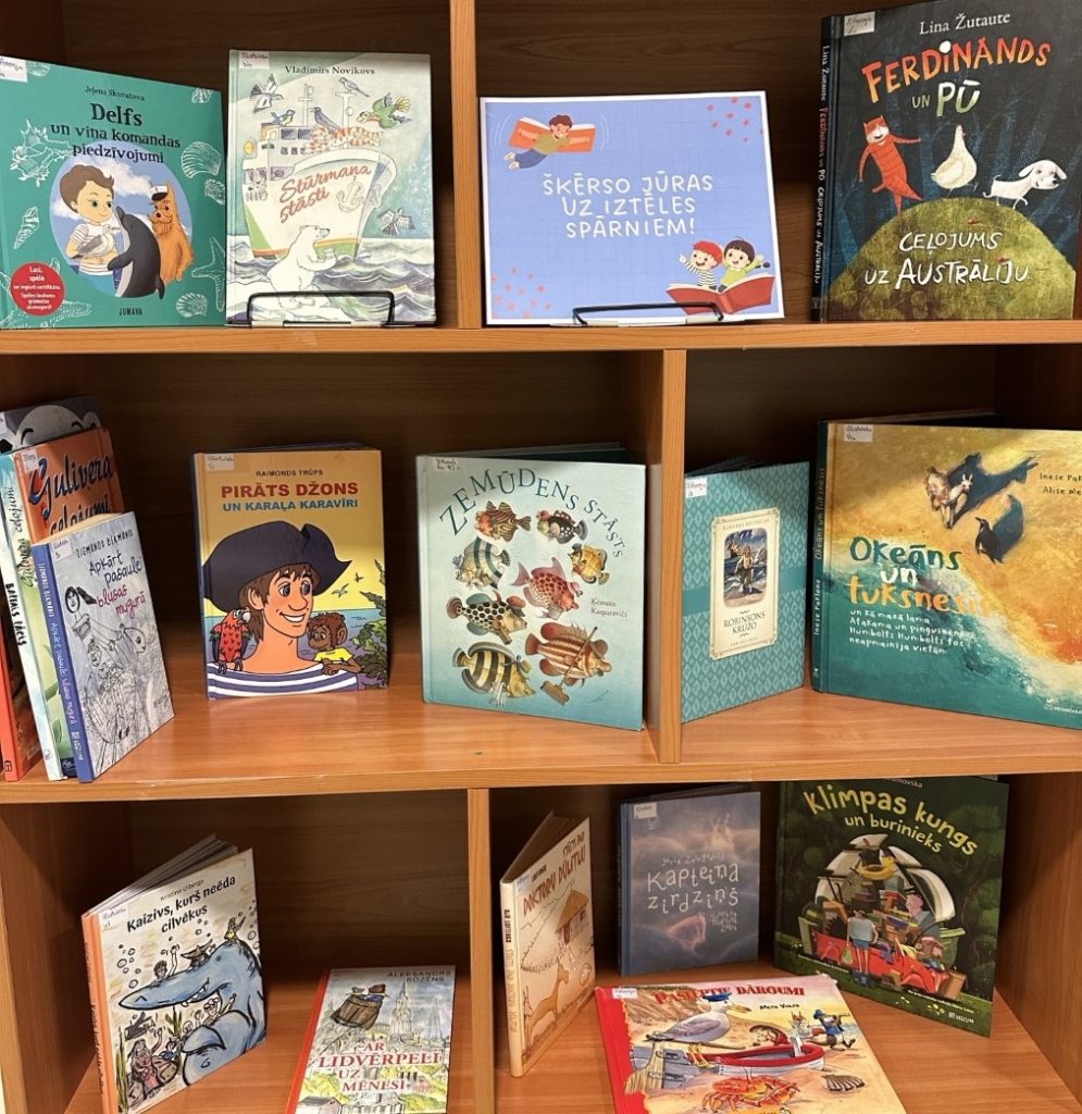 Krāsains foto. Uz koka 3 rindu plauktā saliktās dažādas bērn grāmatas. Uz krāsaina papīra uzraksts - "Šķērso jūras uz iztēles spārniem!"