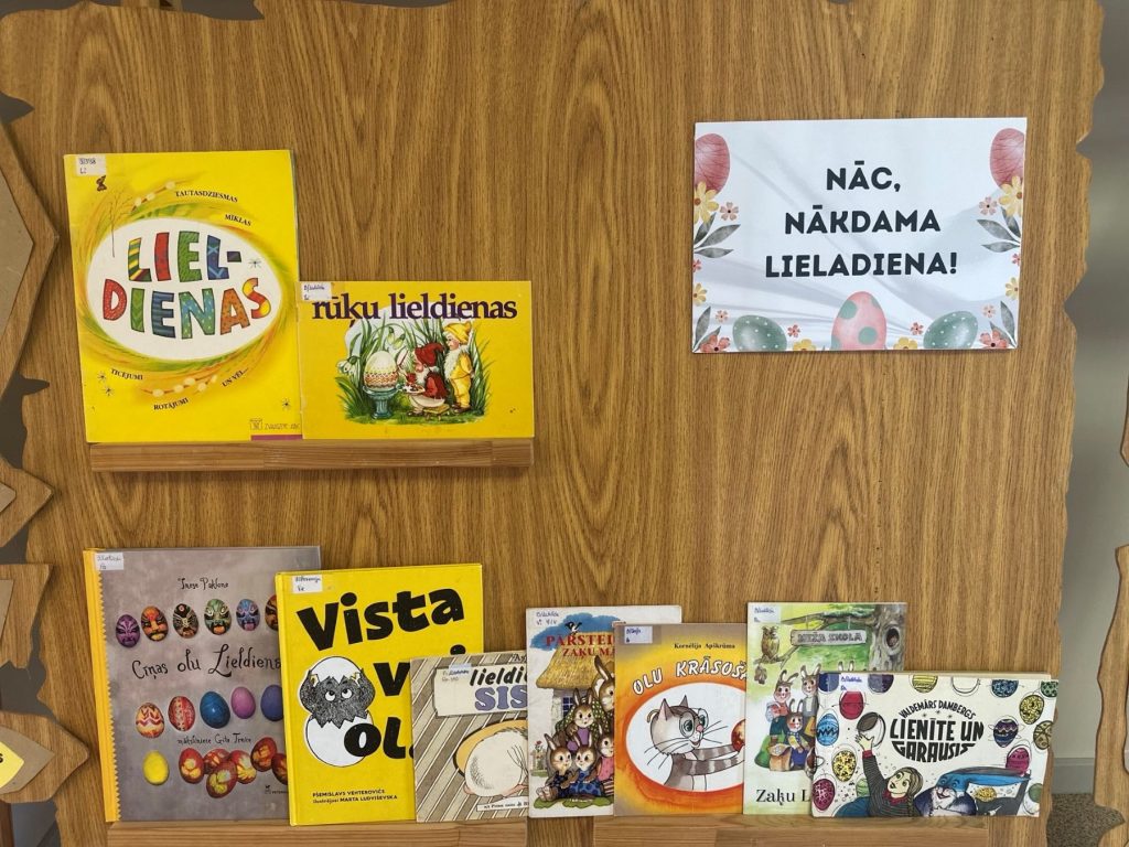 Grāmatu izstāde ar nosaukumu “Nāc, nākdama Lieldiena!”. Uz koka plaukta izkārtotas krāsainas bērnu grāmatas par Lieldienu tematiku – olām, zaķiem un pavasari. Blakus izvietots svētku nosaukuma plakāts ar Lieldienu dekorācijām.
