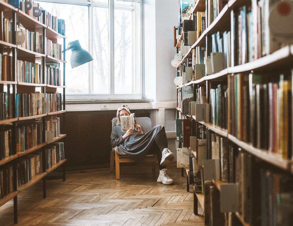 Bibliotēkas telpa, ar augstiem grāmatu plauktiem abās pusēs. Dabiskais gaisma iekrīt no liela loga pa vidu, apgaismojot koka grīdu un radot siltu atmosfēru. Centrā attēlā ir sieviete, sēžot krēslā un lasot grāmatu, pārliekot kājas. Sieviete ģērbusies ikdienišķi un valkā sporta apavus.