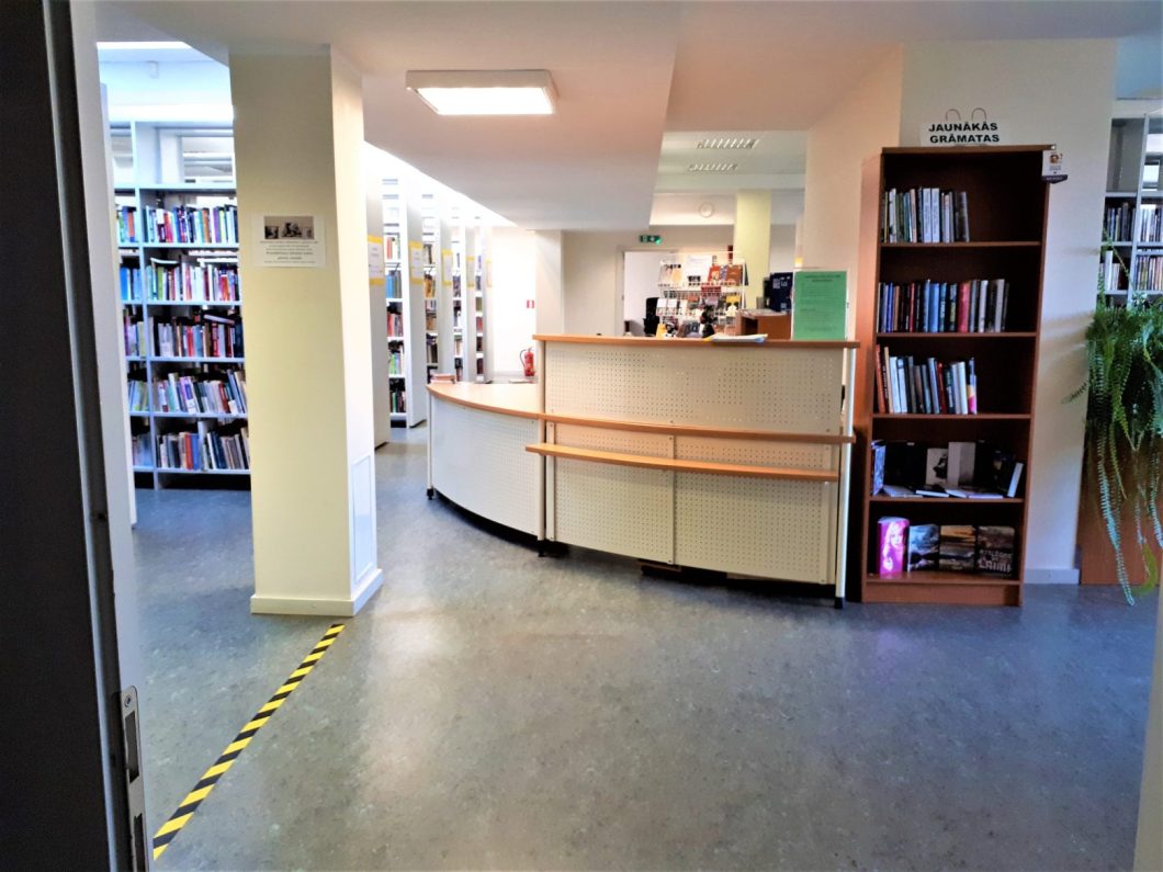 Fotoattēls bibliotēkas iekštelpas. Ir pusapļa pieņemšanas galds, fona augsti plaukti ir grāmatām, un ir atvērta durvju aile, kas ved uz šo zonu. Grīdā ir dzeltena un melna ar dzelteno piesardzības lente, kas norāda uz noteiktu ceļu vai vietu, kur jāizturas piesardzīgi.