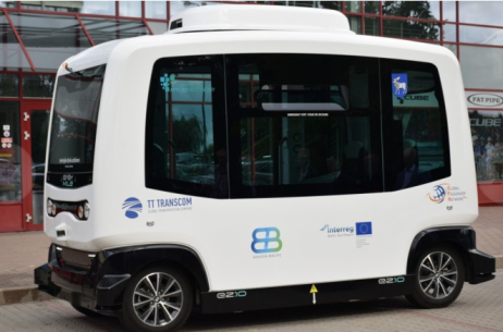 Mazs, baltas krāsas autobuss ar melnu apdari. Uz transportlīdzekļa ir vairāki logotipi un nosaukumi, tostarp “TT TRANSCOM” un “GACHA”. Autobusam ir lielas logu platnes, un šķiet, ka ir aprīkots ar sensoriem vai kamerām augšpusē un sānos, kas, visticamāk, tiek izmantoti navigācijai bez cilvēka vadītāja. Fons ietver sarkano ēku ar lieliem logiem