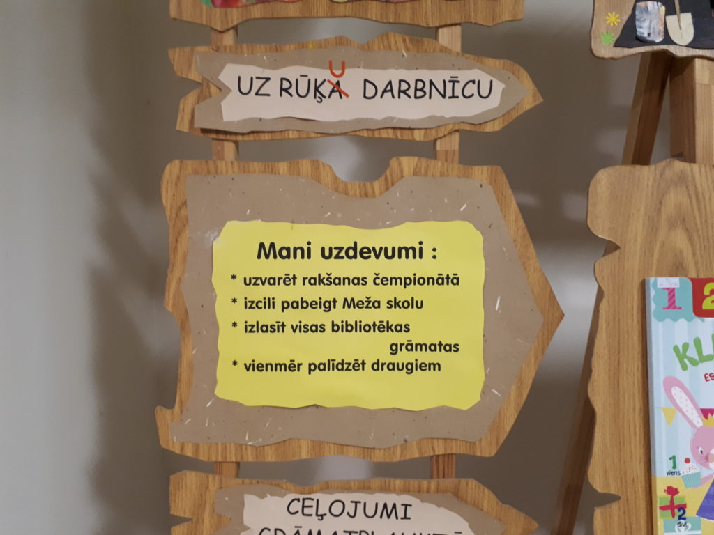 Daļa no koka statīva ar uzrakstiem "Uz Rūķu darbnīcu", "Mani uzdevumi", "Ceļojumi"