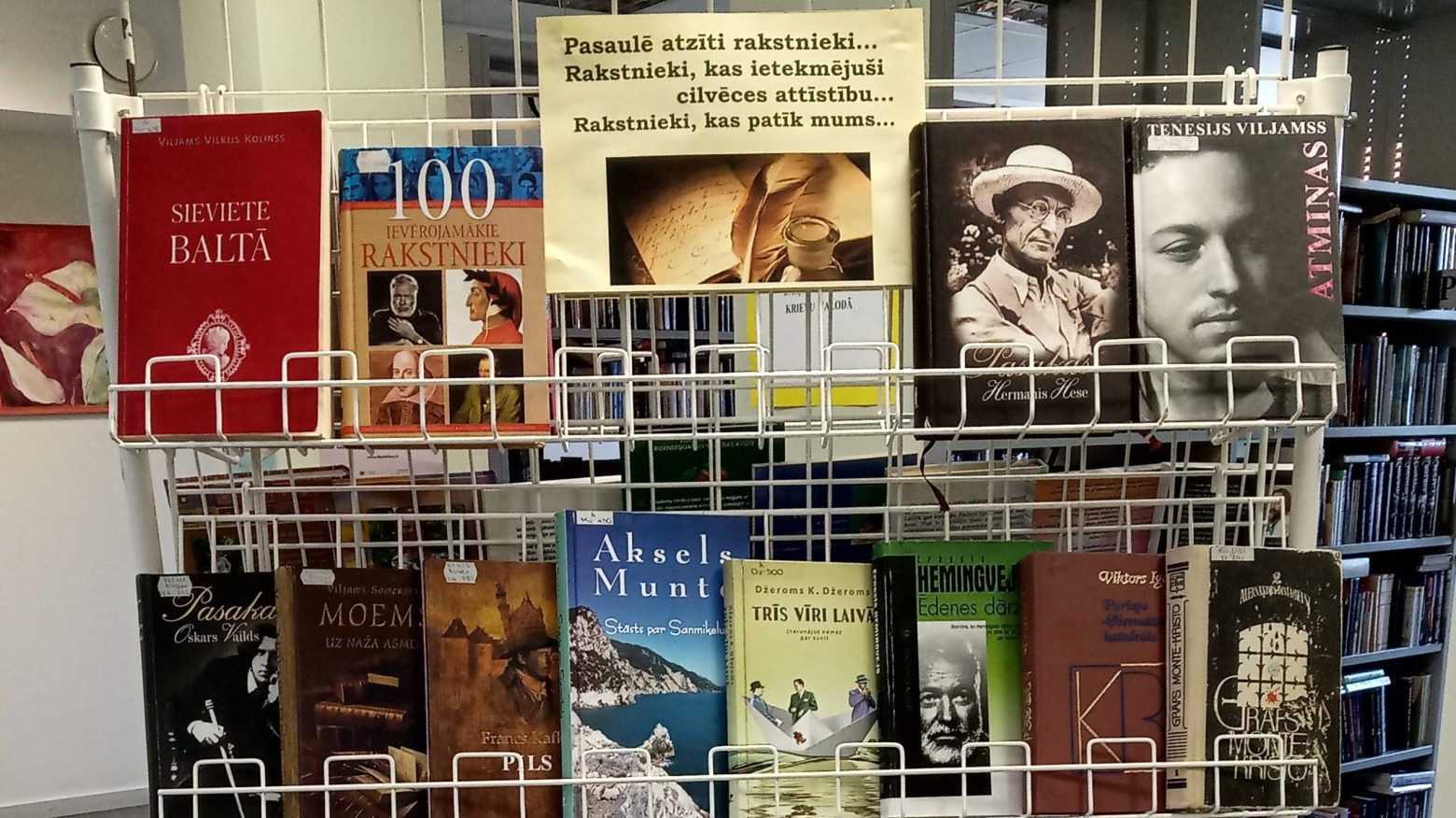 Uz balta, dzelzs stelāžā, bibliotēkas telpā, izliktās vairākas, dažādas grāmatas. Ir uzraksts -"Pasaulē atzīti rakstnieki..."