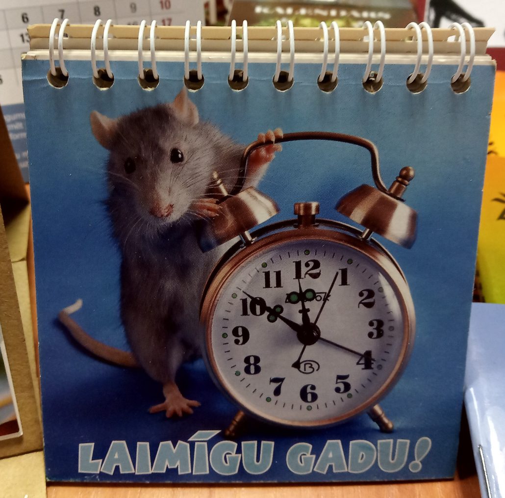 Attēlā redzams kalendārs ar žurkas fotogrāfiju, kas tur modinātājpulksteni. Uz kalendāra ir uzraksts “LAIMĪGU GADU!”