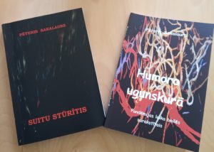 divas grāmatas uz koka virsmas. Kreisajā pusē ir grāmata ar melnu vāku, uz kuras rakstīts “Suitų Stūrītis”. Labajā pusē ir grāmata ar krāsainu abstraktu dizainu uz vāka, nosaukums “Humora ugunskurā”.