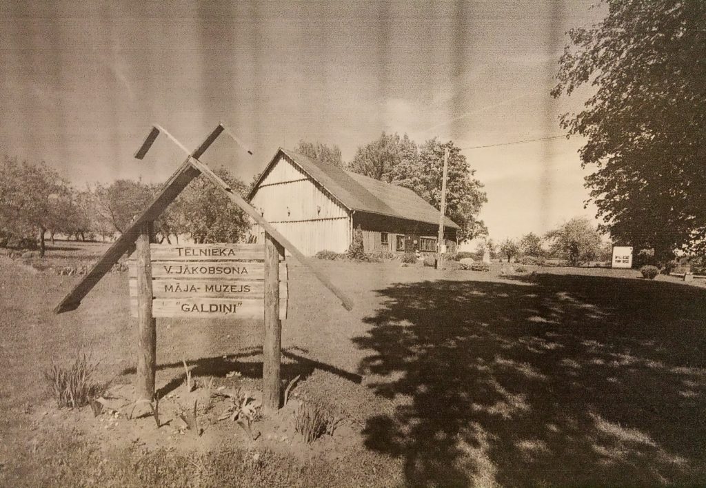 melnbalta fotogrāfija ar lauku ainavu. Priekšplānā ir koka ceļazīme ar uzrakstiem, kas norāda uz fonā esošu ēku, iespējams, muzeju. Apkārt ēkai ir koki, un fotogrāfijai ir vēsturisks izskats.