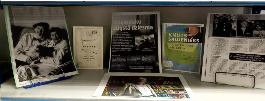 grāmatu un drukātu lapu kolekcija, kas sakārtota vertikāli uz plaukta. Teksts tiem ir latviešu valodā. Lapas ievietotās mazos statīvos vertikāli. Uz lapām melnbalti fotoattēli.