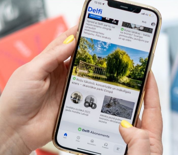 rokas tur viedtālruni. Viedtālrunī ir atvērta lietotne "Delfi," kas ir ziņu portāls. Ekrānā redzami vairāki raksti ar attēliem un virsrakstiem.