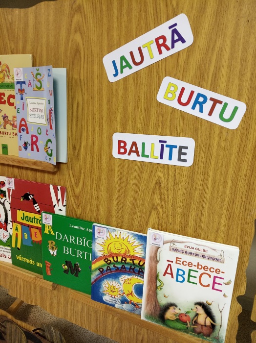 koka virsma, uz kuras ir izvietotas vairākas bērnu grāmatas un krāsaini uzraksti: "JAUTRĀ", "BURTU", "BALLĪTE". Grāmatas ir saistītas ar burtiem un alfabētu, piemēram, "Ece-bece ĀBECE"