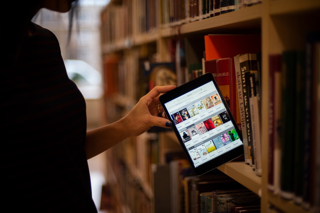 Pie lielā bibliotēkas grāmatu plaukta, stāv sieviešu stāvs melnā, un, it kā, izvelk no plaukta planšeti ar iedegto ekrānu