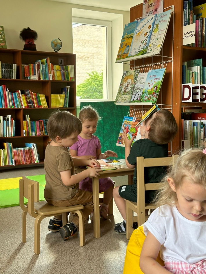 Četri mazie bērni bibliotēkā, sēžot pie galda un skatās grāmatas. Grāmatu plaukti ir pilni ar dažādām bērnu grāmatām.