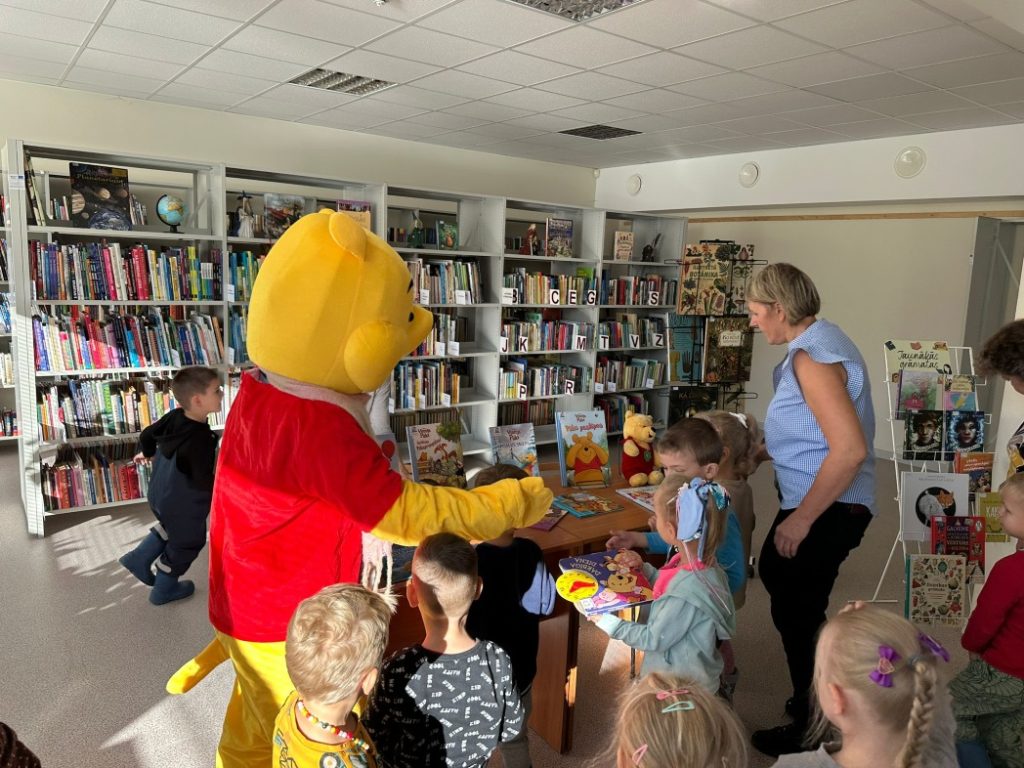 bibliotēka ar grāmatu plauktiem fonā. Priekšplānā ir cilvēks, kurš ir tērpies kā Vinnijs Pūks, un apkārt viņam ir bērni. Uz galda ir dažādas grāmatas un rotaļlietas, kas saistītas ar šo varoni. Pieaugušais, iespējams, skolotājs vai bibliotekārs, palīdz bērniem. 