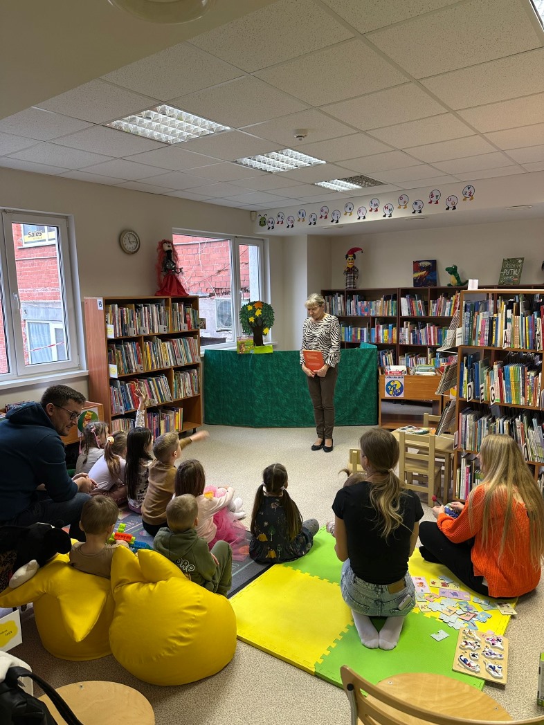 bērnu bibliotēka, notiek pasākums bērniem. Telpā ir daudz grāmatu plauktu, kas piepildīti ar grāmatām. Priekšplānā ir vairāki bērni, kas sēž uz spilveniem un paklājiem, klausoties pieaugušo, kas stāv priekšā. Pieaugušais tur rokās grāmatu. Telpā ir arī dažas rotaļlietas un puzles, kas izklātas uz grīdas. Pie sienām ir dekorācijas un zīmējumi