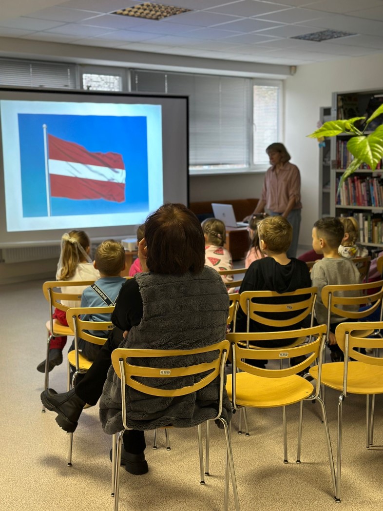 telpā, uz baltās tāfeles projicētā Latvijas karoga bilde. tai priekšā rindās uz krēsliem sēž bērni un daži pieauguši. Redzami grāmatas plaukti.