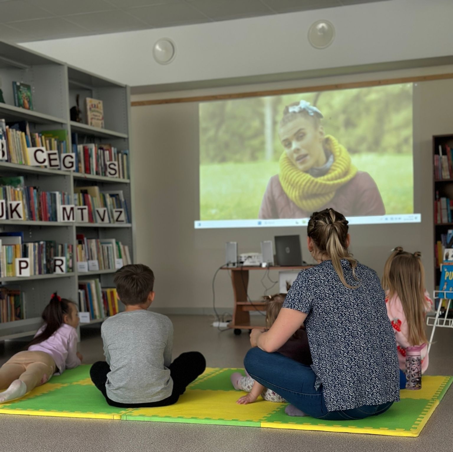 Bērni, kas sēž uz grīdas krāsaina tepiķī un skatās video, kas tiek rādīta uz projektora ekrāna bibliotēkā. Fonā redzama grāmatu plaukti.