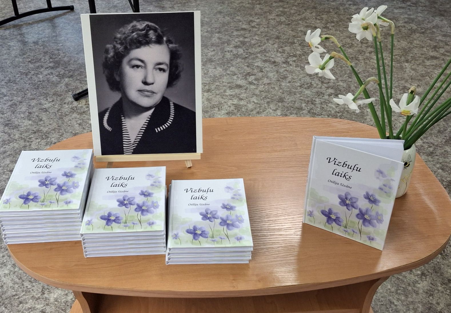Koka ovālais galdiņš ar grāmatām "Vizbuļu laiks", kuru autore ir Ottilija Uzkāne. Uz galda atrodas arī melnbalta autores fotogrāfija un balti ziedi vāzē. Visticamāk, attēlots piemiņas vai grāmatas atvēršanas pasākums.
