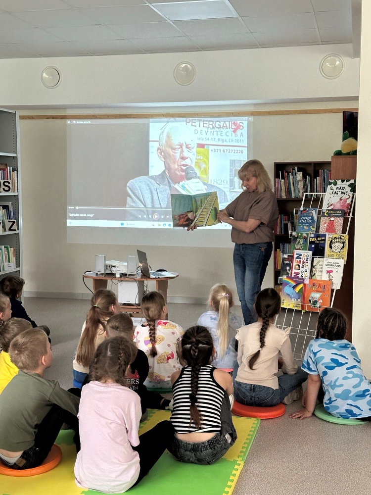 Sieviete, kas rāda bērnu grāmatu grupai bērnu bibliotēkā. Bērni sēž uz krāsainā paklājā. Fonā redzams projektors ar attēlu uz ekrāna – tur redzams vecāks vīrietis, rakstnieks, teksts "PĒTERGAILS" un kontaktinformācija. Blakus redzama grāmatu izstāde vai stends ar bērnu literatūru.