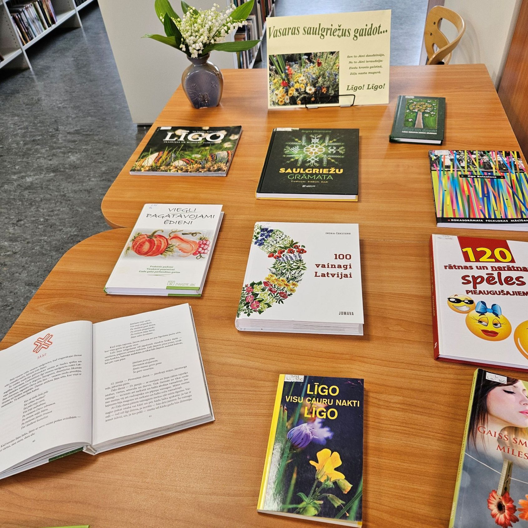 Grāmatu izstāde bibliotēkā, kas veltīta vasaras saulgriežu svētkiem – Līgo un Jāņiem. Grāmatas ir izkārtotas uz koka galda. Galda centrā redzams arī uzraksts "Vasaras saulgriežus gaidot... Līgo! Līgo!". Dekorācijai izmantota ziedu vāze ar maijpuķītēm.