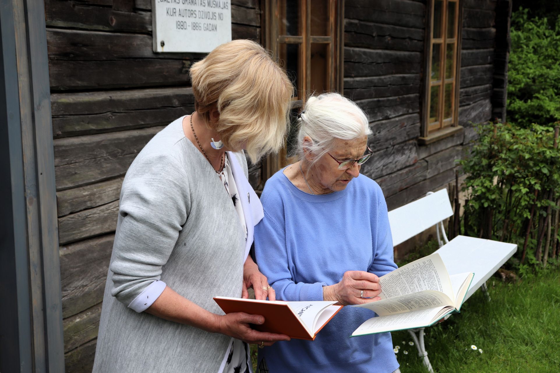 Attēlā redzami divi vecāka gadagājuma sievietes, kas stāv pie vecas koka ēkas. Viena sieviete tur atvērtu grāmatu un šķiet, ka lasa vai apspriež tās saturu ar otru cilvēku, kurš arī tur grāmatu. Fonā redzams balts sols un zaļumi.