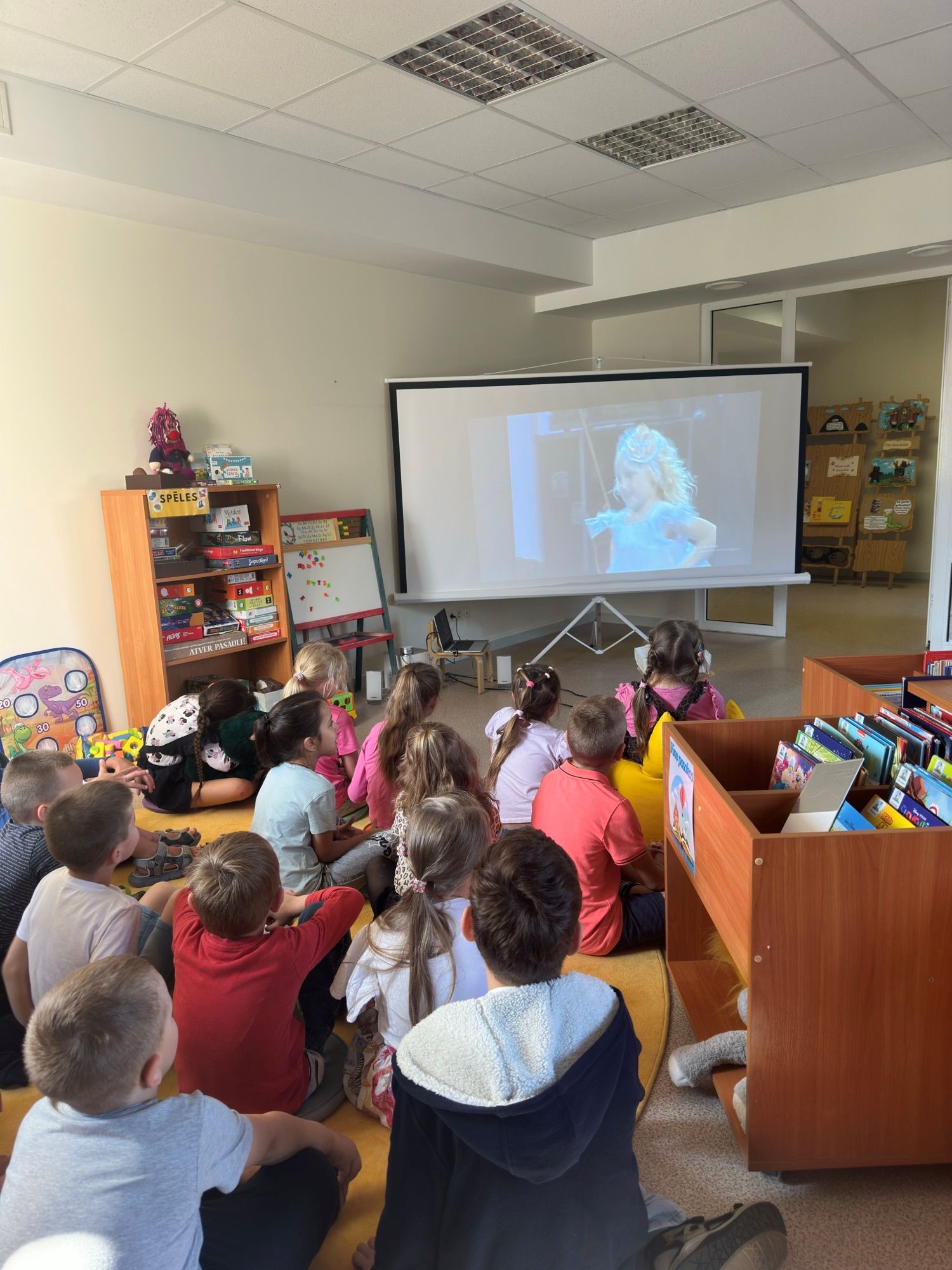 Bērni, kas sēž uz grīdas un skatās video projekciju, kurā redzama meitene zilā kleitā. Telpā ir plaukti ar spēlēm un grāmatām.