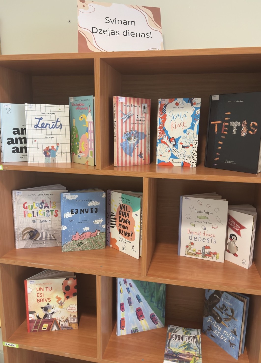 Grāmatu izstāde bibliotēkā ar uzrakstu “Svinam Dzejas dienas!”. Koka Plauktos izvietotas bērniem un jauniešiem domātas grāmatas – dzejoļu krājumi un citi literāri darbi, piemēram, “Zenīts”, “Skolā klase”, “Tētis” u.c.