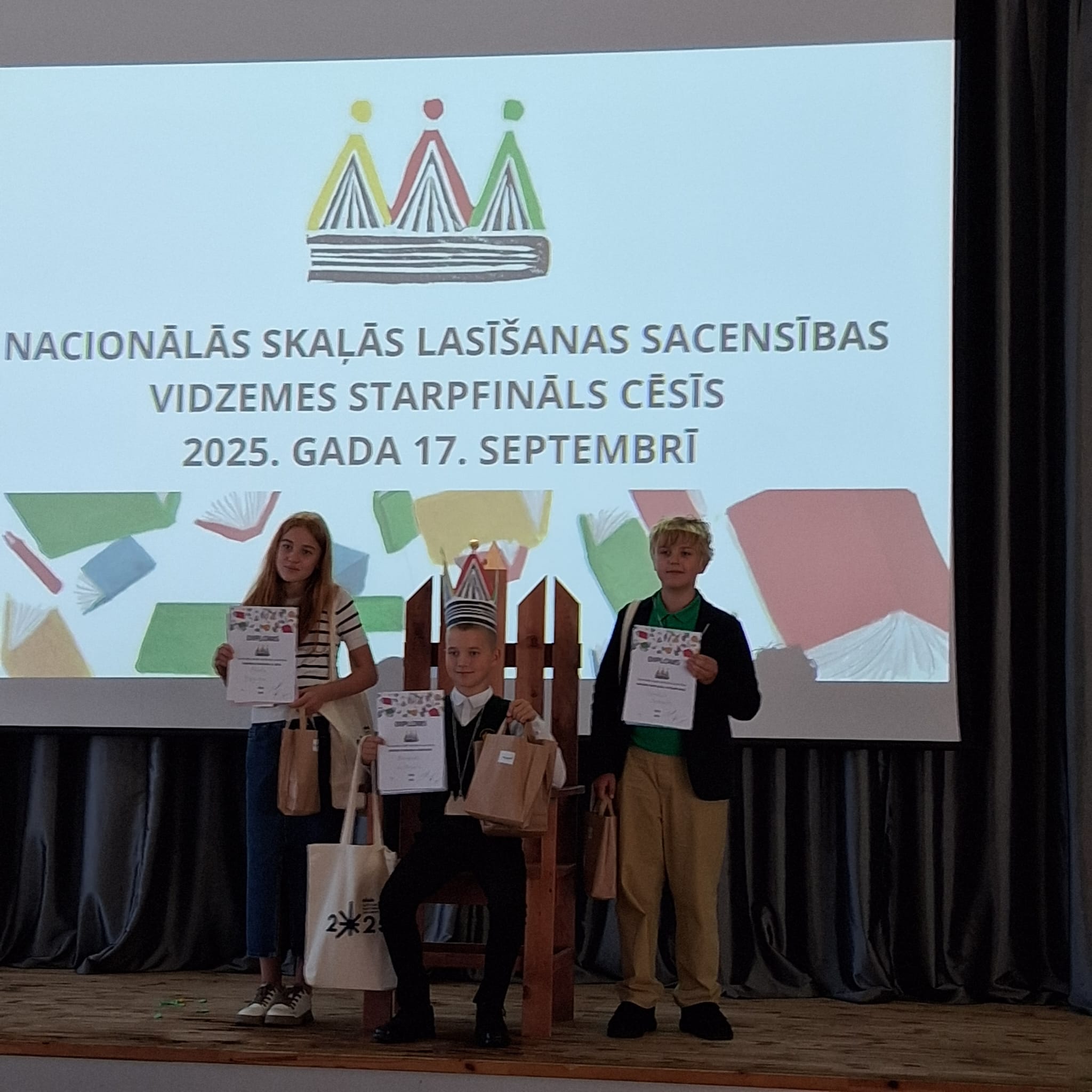Trīs bērni uz skatuves ar diplomiem un balvām, aiz viņiem ekrānā redzams uzraksts par Nacionālās skaļās lasīšanas sacensībām Vidzemes starpfinālā Cēsīs, 2025. gada 17. septembrī. Vidū esošais bērns sēž krēslā ar papīra kroni galvā.