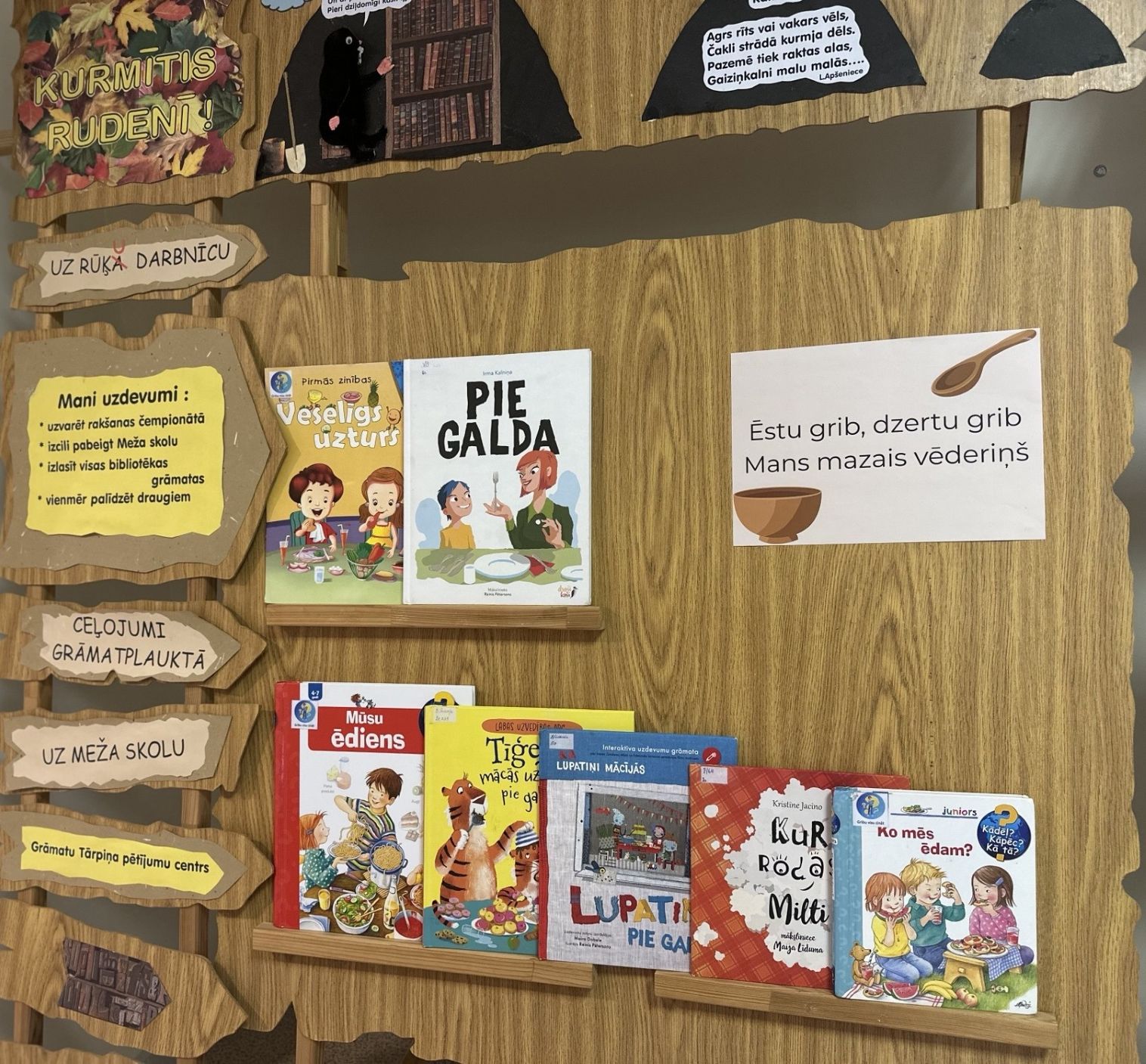 Bibliotēkas bērnu grāmatu izstāde par veselīgu uzturu un ēšanas paradumiem. Plauktā izkārtotas grāmatas ar nosaukumiem “Veselīgs uzturs”, “Pie galda”, “Ko mēs ēdam?” u.c., bet dekorācijās izmantoti rudens un meža tēli ar uzrakstiem un dzejolīti par ēšanu.