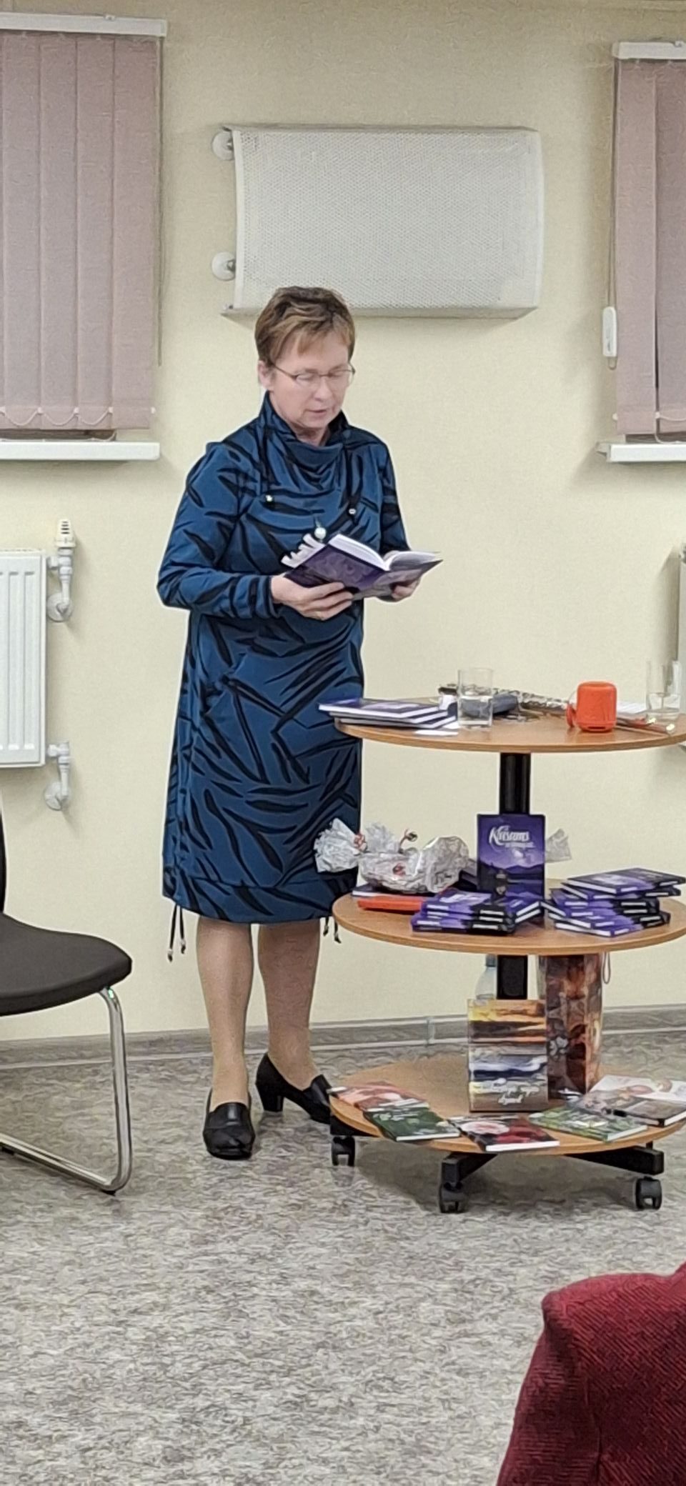 Sieviete, kas stāv pie galdiņa un lasa no grāmatas vai burtnīcas. Uz galdiņa novietotas vairākas grāmatas, papīri un nelieli priekšmeti, kas norāda uz literāru vai radošu pasākumu.