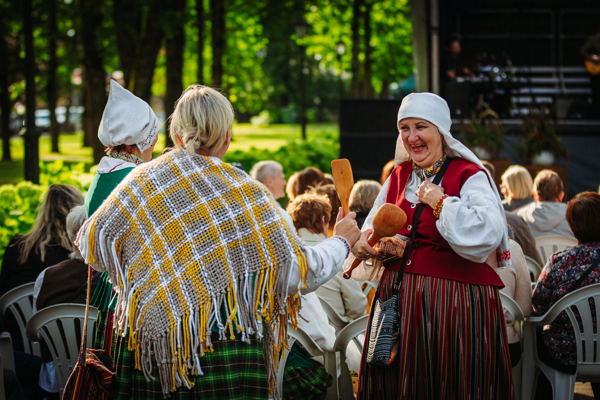 Brīvdabas pasākums, kurā piedalās cilvēki tautas tērpos. Priekšplānā divas personas ir ģērbtas tradicionālajos latviešu tautas tērpos – rakstainās villaines, svītraini brunči, lina krekli un galvassegas. Viņas rokās tur koka priekšmetus, kas izskatās pēc tradicionāliem mūzikas instrumentiem vai ritma rīkiem. Fonā sēž skatītāji uz baltām plastmasas krēsliem, bet tālāk redzama skatuve ar mūzikas aparatūru. Apkārt ir zaļi koki un saulaina diena, kas rada svētku un kultūras notikuma noskaņu.
