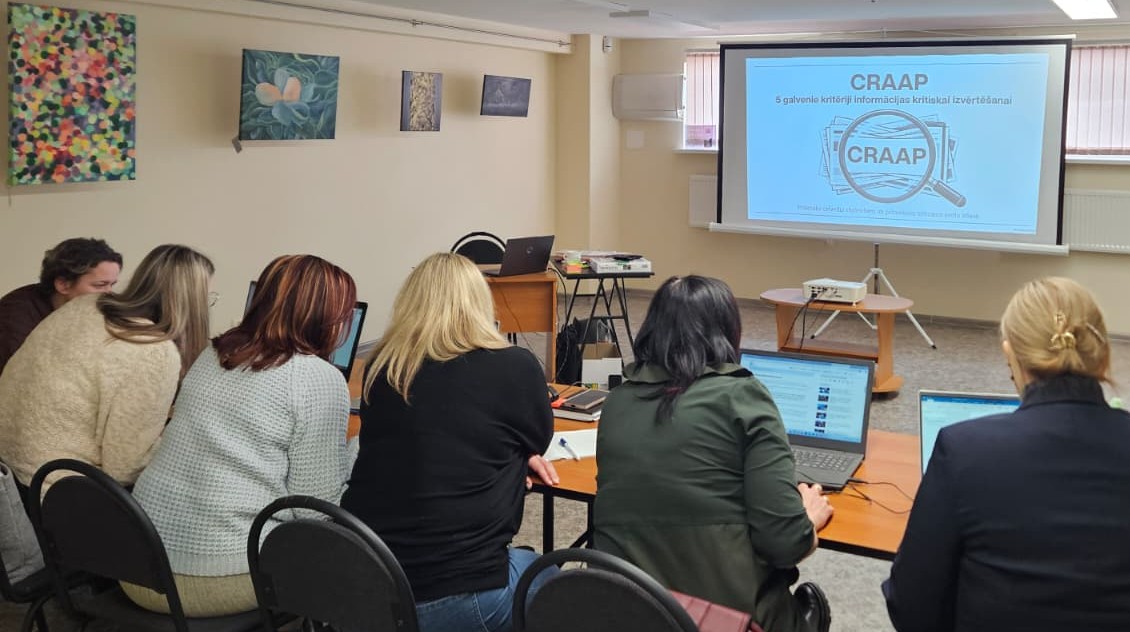 Mācību nodarbība vai seminārs. Dalībnieki sēž pie galdiem ar klēpjdatoriem un skatās prezentāciju uz ekrāna par informācijas kritisku izvērtēšanu (CRAAP kritēriji).