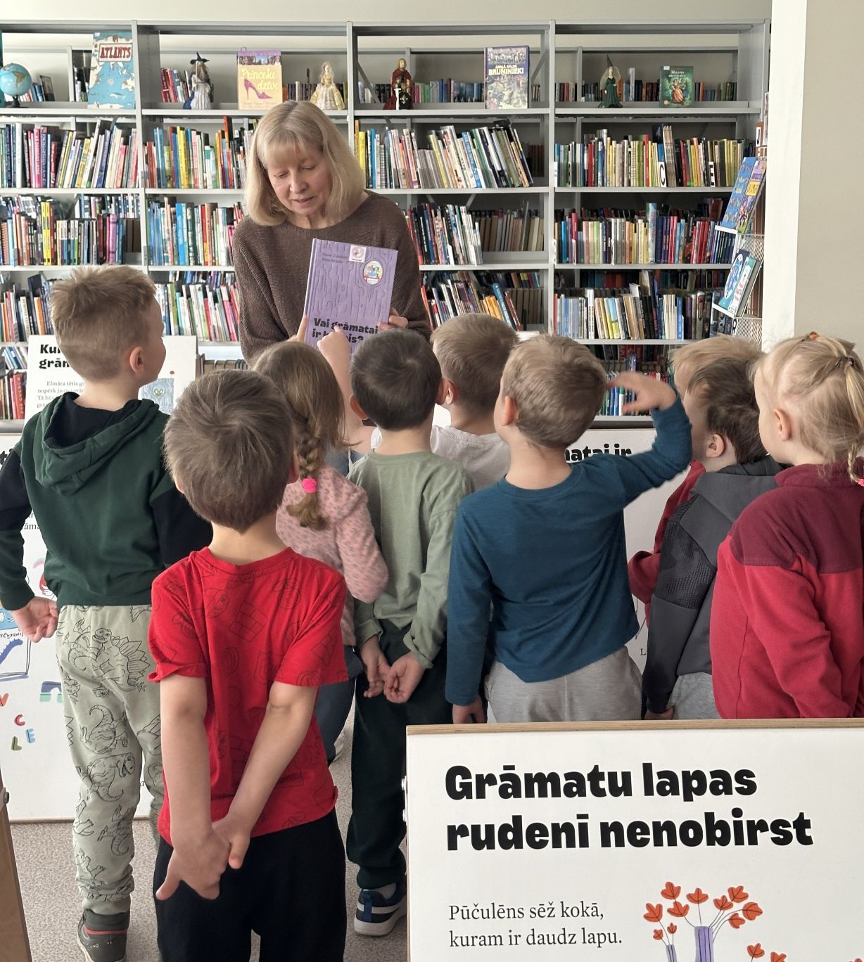 Sieviete (iespējams, skolotāja vai bibliotekāre), kas bibliotēkā rāda grāmatu mazai bērnu grupai. Bērni stāv apkārt un uzmanīgi skatās, fonā redzami grāmatu plaukti.