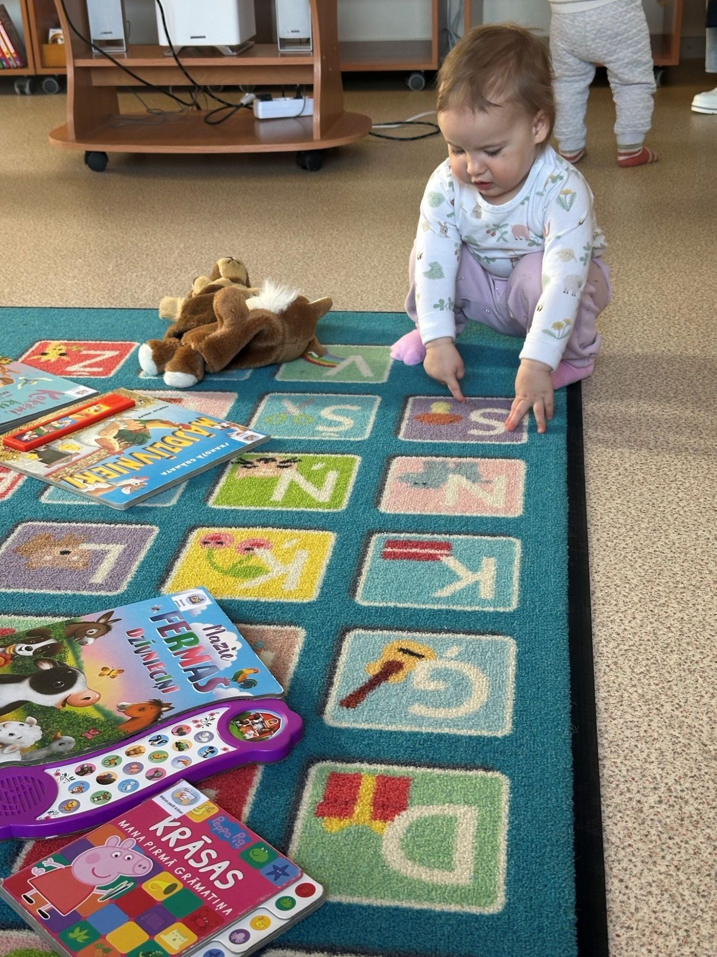 Mazs bērns sēž uz paklāja ar burtiem, skatās grāmatas un ar pirkstiem rāda uz burtiem.