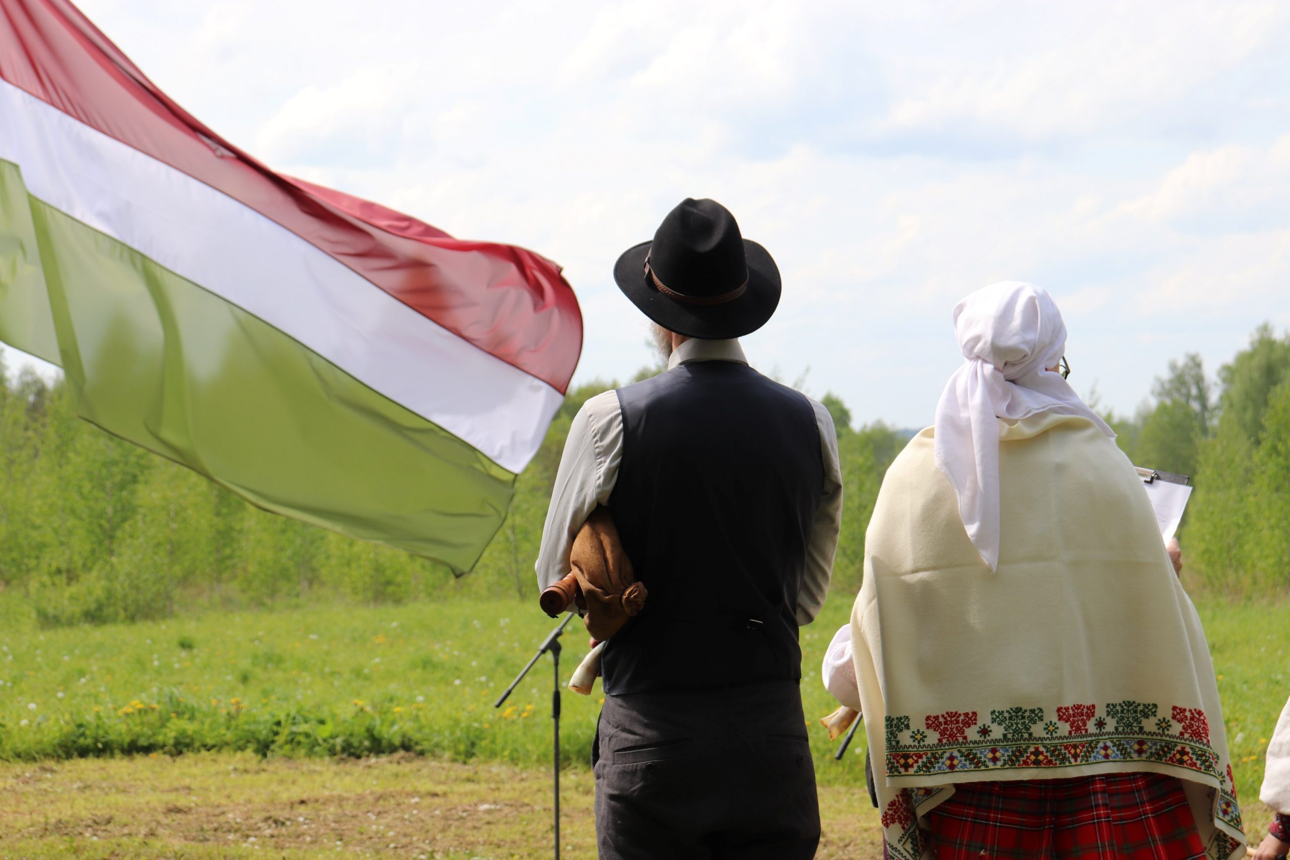 Divi cilvēki tautastērpos stāv pļavā, skatoties uz karogu, kas plīvo vējā. Blakus redzams mikrofons, iespējams, notiek kāds pasākums vai svētki. Karoga krāsas: sarkans, balts, zaļš.