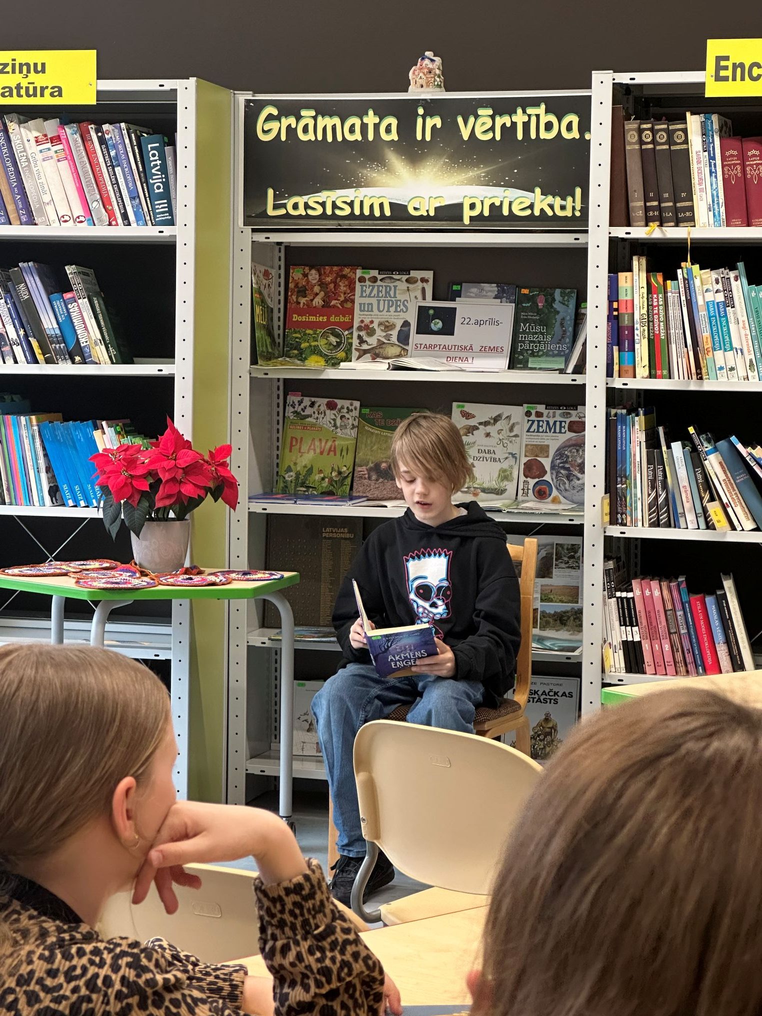 Skolēns-zēns bibliotēkā, kurš sēž un lasa grāmatu citu bērnu priekšā. Fonā ir grāmatu plaukti un uzraksts par lasīšanu.