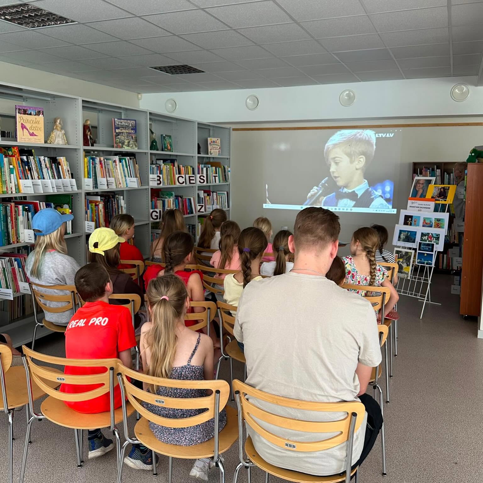 Bērni bibliotēkā, sēž uz krēsliem un skatās video projekciju uz sienas. Sānos arī lieli grāmatu plaukti.
