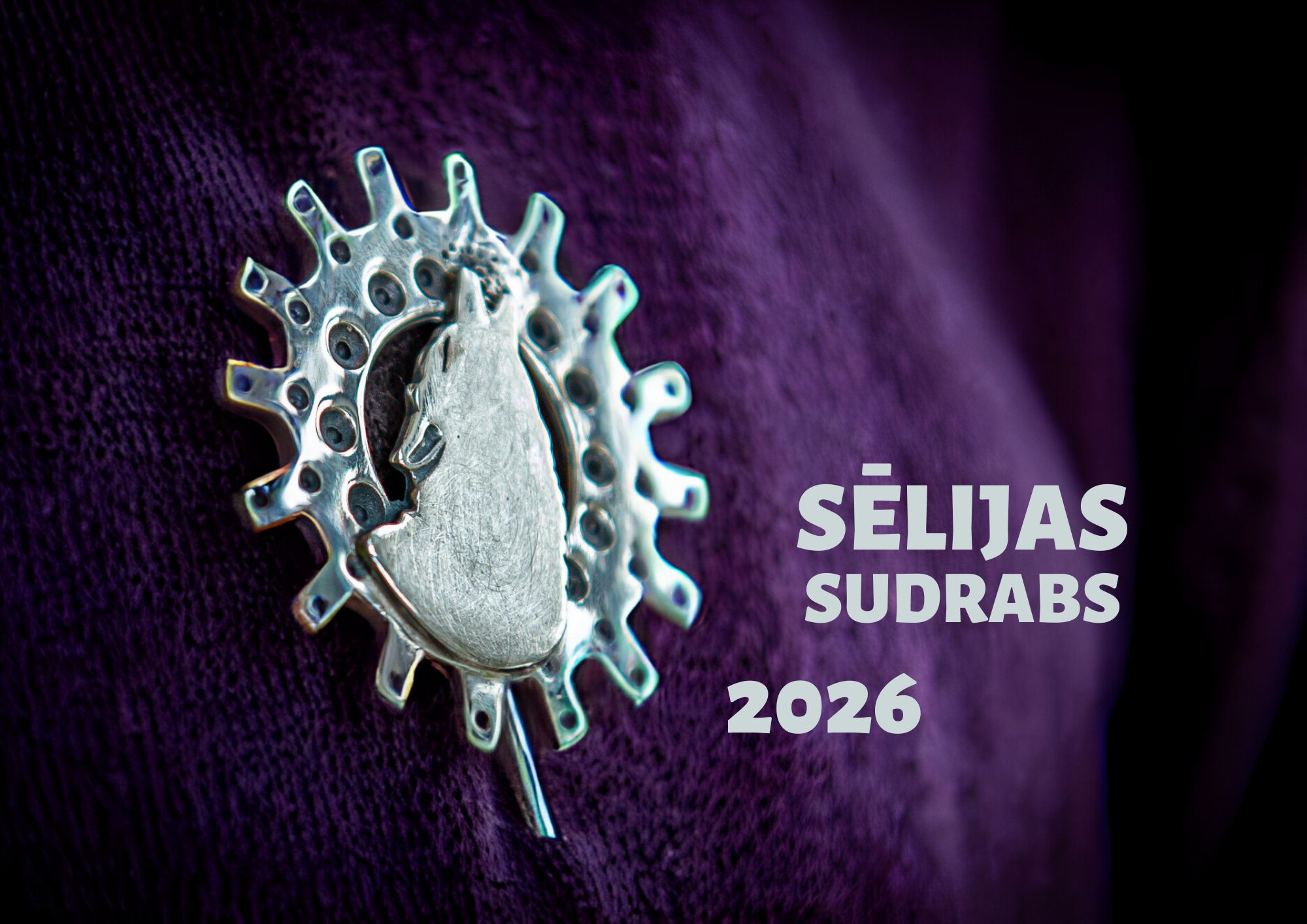 Sudraba piespraude ar stilizētu suņa galvas figūru saules formas ornamentā. Piespraude piestiprināta pie tumši violeta auduma, attēla labajā pusē redzams uzraksts "SĒLIJAS SUDRABS 2026".