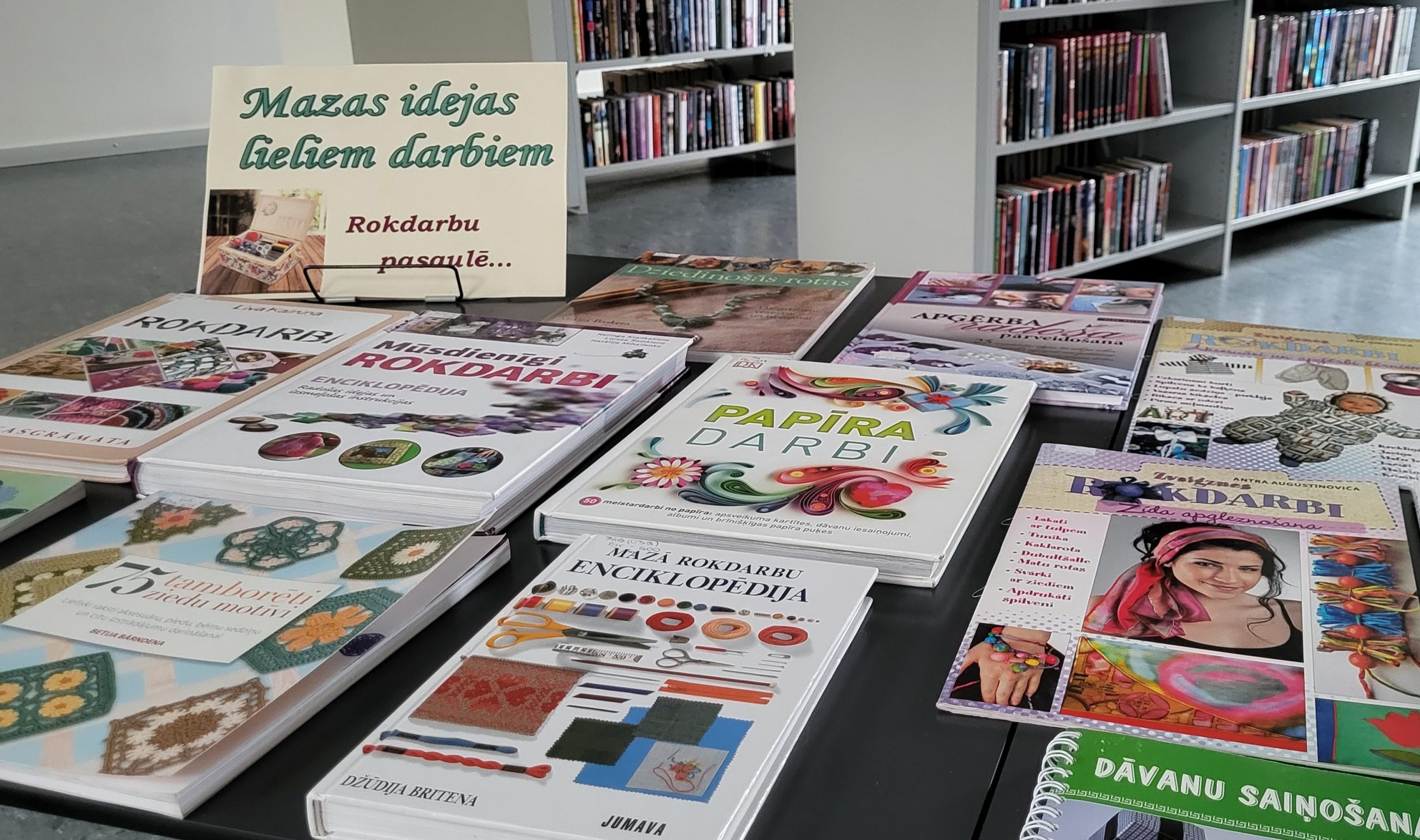 Uz galda saliktas rokdarbu grāmatas, blakus grāmatām uzlikta A4 lapa ar nosaukumu ''Mazas idejas lieliem darbiem'', kas ir izstādes nosaukums.